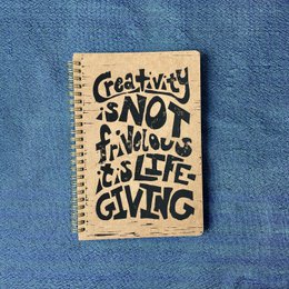 A spiral-bound notebook with the text "Creativity is not frivolous, it's life-giving" handwritten on the cover. The notebook has blank pages inside.
