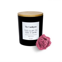 A tall, round black candle with a white label that reads "Too Glam To Give A Damn". The candle is surrounded by red rose petals and has a bamboo lid. The candle appears to be a scented candle based on the product title.