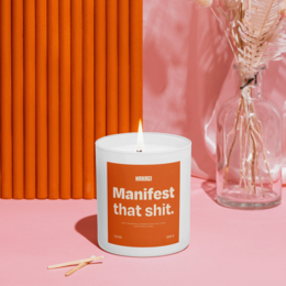 A round, tall, orange-colored scented candle with the text "Manifest that shit" printed on the label. The candle is surrounded by a pink and orange background, creating a cozy and vibrant atmosphere.