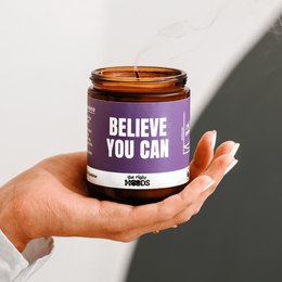A scented, handmade soy wax candle in a glass jar with the text "BELIEVE YOU CAN" printed on the label. The candle has an amber-colored flame and is surrounded by a purple gift box.
