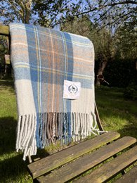 A large, soft, and cozy tartan blanket in muted blue and beige tones, made from 100% recycled wool. The blanket has a classic plaid pattern and fringed edges, making it a perfect addition to any living room or bedroom.