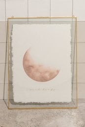A poster featuring a soft, pastel-colored crescent moon against a light background, framed in a simple wooden frame.
