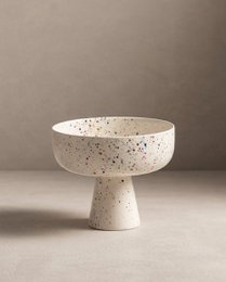  A round, white ceramic bowl with a pedestal base, featuring a speckled pattern of colorful dots throughout.