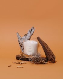 A tall, round white candle with a wooden wick, surrounded by natural elements like twigs and stones, creating a rustic and earthy aesthetic.