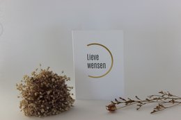 A greeting card with the text "Liebe wünscht" (Wishes love) and a simple circular design. The card is surrounded by dried flowers and branches, creating a natural and minimalist aesthetic.