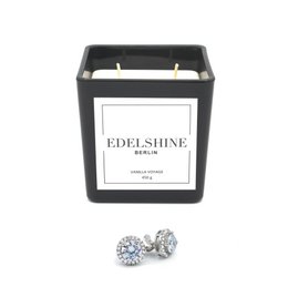 Large scented candle with classic diamond stud earrings. The candle has a black container and the earrings feature two round, sparkling diamonds.