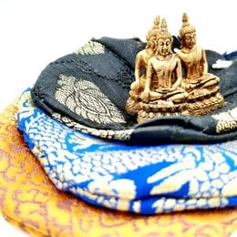 A small golden Buddha statue sitting on a colorful, patterned cushion. The cushion has a dark background with intricate designs in blue, orange, and white.