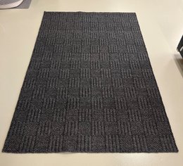 A large, rectangular area rug in a dark charcoal gray color. The rug has a textured, woven pattern with vertical stripes. The rug is suitable for use in living rooms, bedrooms, or other indoor spaces.