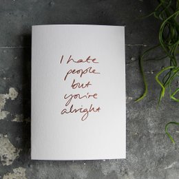 A hand-foiled greeting card with the text "I hate people but you're alright" written in red cursive font on a white background.