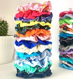 A colorful stack of eco-friendly scrunchies in various vibrant colors and patterns, including solid colors, stripes, and floral designs. The scrunchies appear to be made of soft, stretchy fabric and are neatly stacked, showcasing the variety of options available.