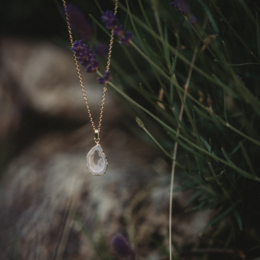 Agate geode necklace with a teardrop-shaped pendant on a delicate chain, surrounded by lush purple flowers.