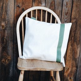 Linha cushion, a decorative pillow with a white fabric cover and contrasting leather trim in green, burgundy, or lamb leather. The cushion has a simple, minimalist design that would complement a variety of home decor styles.