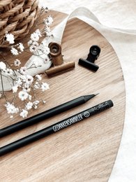 Two black pencils with the brand name "Schulkind" printed on them, surrounded by white daisy flowers on a wooden surface.