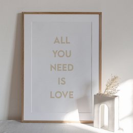 This wall art features the text "ALL YOU NEED IS LOVE" in a minimalist, typographic design. The text is displayed in a neutral, light color against a plain background, creating a simple and elegant aesthetic. The product is available in multiple size variants, making it suitable for various wall spaces.