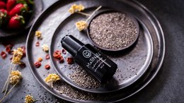 A dark, round facial oil serum in a glass bottle on a metal tray, surrounded by red berries and yellow flowers.