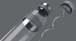 A tall, cylindrical glass carafe with a light grey silicone band around the middle. The carafe has a clear glass body that allows the contents to be visible, and a silver-colored metal lid on top.