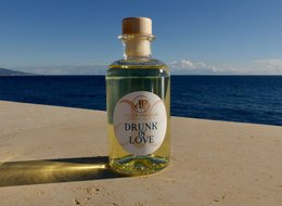 Reed diffuser with "Drunk in Love" label, glass bottle with amber-colored liquid, wooden reed sticks, and a beach setting with blue ocean in the background.