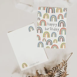 The birthday card features a colorful rainbow pattern with the text "happy birthday" printed on it. The card is presented on a wooden surface with a beige fabric and some natural elements, creating a warm and inviting atmosphere.