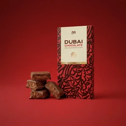 Milk chocolate bar with speculoos and kunafa flavors, brown color, grid-scored segments, luxury gift packaging with red patterned box.