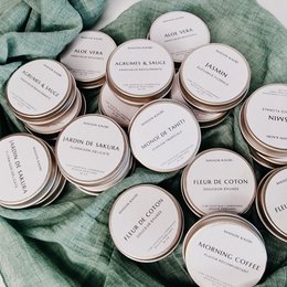 Assorted scented candles in various fragrances including Sweet Lychee, Fleur de coton, Jardin de sakura, and more. The candles are presented in round white containers on a green fabric background.
