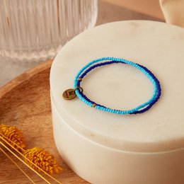  A colorful beaded bracelet with a metal charm that says "#WITHREFUGEES". The bracelet is made up of blue and turquoise beads arranged in a repeating pattern.