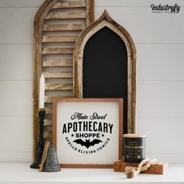 Rustic wooden sign with "Main Street Apothecary No1" text, featuring an arched frame and a black chalkboard background. The sign is surrounded by decorative black bat silhouettes and small pumpkins, creating a cozy, autumn-inspired atmosphere.