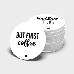 Round, white coasters with the text "BUT FIRST COFFEE" printed on them. The coasters are stacked and displayed in a brown cardboard container.