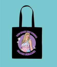 Black tote bag with a graphic illustration of a blonde woman in a pink dress. The text on the bag reads "Taylor Swift Shimmer Tote Bag" and includes a circular design element.