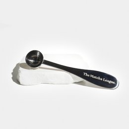 Stainless steel matcha tea scoop with a dark wooden handle. The scoop is resting on a pile of matcha powder, showcasing its intended use for measuring and serving matcha tea.