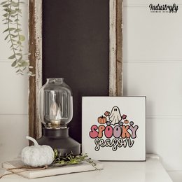 A small, square painting featuring a spooky ghost design in black, orange, and red colors. The painting is displayed on a wooden surface alongside a glass vase with dried flowers.
