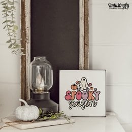 A small, square painting featuring a spooky ghost design in black, orange, and red colors. The painting is displayed on a wooden surface alongside a glass vase with dried flowers.