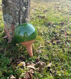 A green, round planter for plants, mounted on a wooden stand and placed in a grassy, forested area.