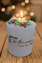A round, white scented candle with a lit wick, surrounded by holly leaves and berries. The candle has the text "Tis the season" printed on it.