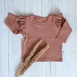 The product is a Ruffle Longsleeve Rib Peach baby shirt. It is made of a peach-colored ribbed material and features ruffled sleeves. The shirt is available in sizes ranging from 56 to 140 cm. This description provides key details about the product's color, material, style, and size options that would be relevant for an ecommerce search.