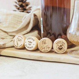 A cork and wood wine bottle stopper with the text "Bonne Fête Papi" and various designs like "Certifié Meilleur Papi", "Chef Papi", "Super Papi", and "Papi Famille Amour Apéro". The stopper also has a stamp that says "Certifié Meilleur Papi du Monde".