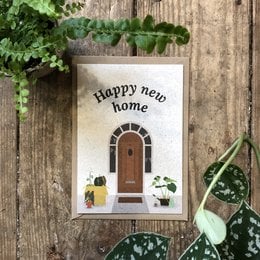 A postcard with the text "Happy new home!" and an illustration of a wooden door with plants around it, set against a wooden background with greenery.