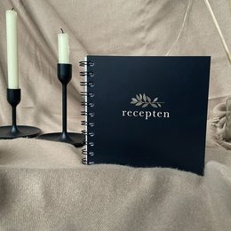A black spiral-bound recipe book with the word "recepten" printed on the cover, accompanied by a pen and a notebook on a wooden surface.