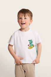 'White cotton t-shirt with a print of a crocodile on a surfboard, suitable for kids aged 18 months to 11 years.'
The product is a white cotton t-shirt with a print of a crocodile on a surfboard, suitable for kids aged 18 months to 11 years. The t-shirt is available in various sizes from 18-24 months (86cm) to 10-11 years (140cm). The print on the t-shirt features a green crocodile riding a yellow surfboard in a wavy, blue background, which gives the impression of a fun and playful design.