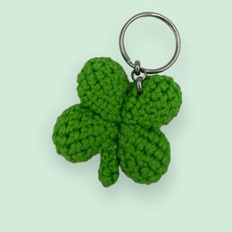 Green crocheted four-leaf clover keychain, made of soft yarn, with a metal ring for attaching to keys or bags.
