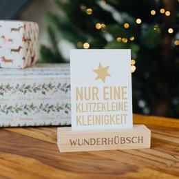 A small, rectangular wooden card holder with the text "Nur eine glitzernde Kleinigkeit" (Only a glittering little thing) and the brand name "Wunderhübsch" printed on it. The card holder is placed on a wooden surface with a festive background.