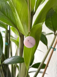 A delicate pink opal pendant on a gold chain, suspended against a backdrop of lush green leaves.