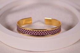 A gold-colored bangle bracelet with a decorative pattern featuring a repeating diamond-shaped design in shades of purple and white.