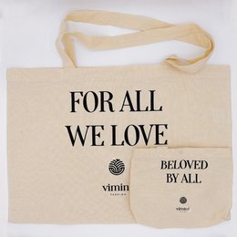 Beige canvas tote bag with black text "FOR ALL WE LOVE BELOVED BY ALL" and a small logo. The bag is being held by a woman and child, suggesting it is a reusable shopping bag or tote.