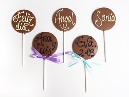 Personalized milk chocolate lollipops with festive text and designs, including "Feliz dia", "Angel", "Santa", "Feliz 2019", and "Amor y paz". The lollipops are presented on sticks and tied with colorful ribbons, making them a delightful holiday or gift item.