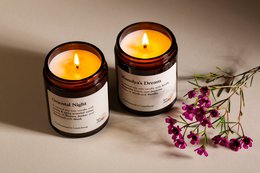 Two round, amber-colored glass candle jars with lit candles. The jars have a label that says "Sol e Mare". The candles are surrounded by purple flowers.