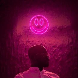  A round, pink neon smiley face lamp on a dark background, with a laptop and other lighting fixtures visible.