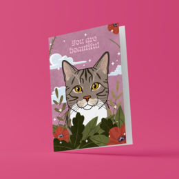 A greeting card featuring a close-up portrait of a tabby cat with green eyes against a pink background with floral accents. The text on the card reads "You are beautiful."