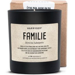 A round, tall, scented candle in a black glass container with the label "HAPPYSOY FAMILIE" printed on it. The candle is surrounded by natural elements like leaves, suggesting it has a natural, earthy fragrance.