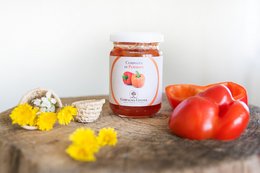Red bell peppers, yellow flowers, and a jar of Composta di Peperoni seasoning on a wooden surface.