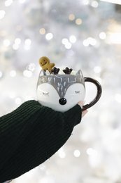 A white ceramic mug with a sleeping deer design, held in a gloved hand against a snowy, bokeh background.